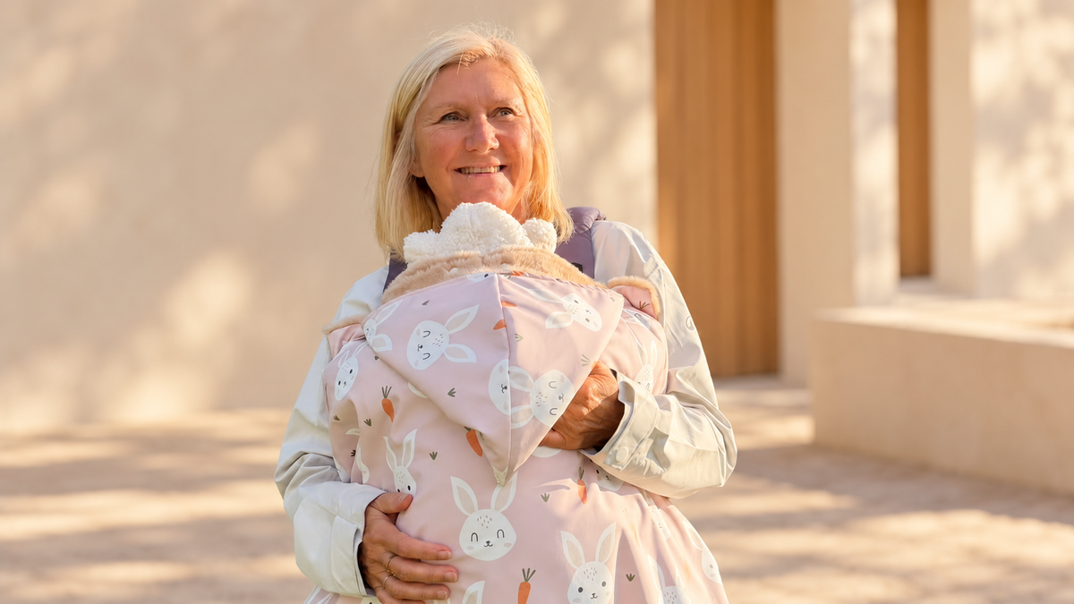Femme souriante portant un bébé contre elle, enveloppé dans une couverture de portage rose à motifs lapins et carottes, doublée de fourrure beige.