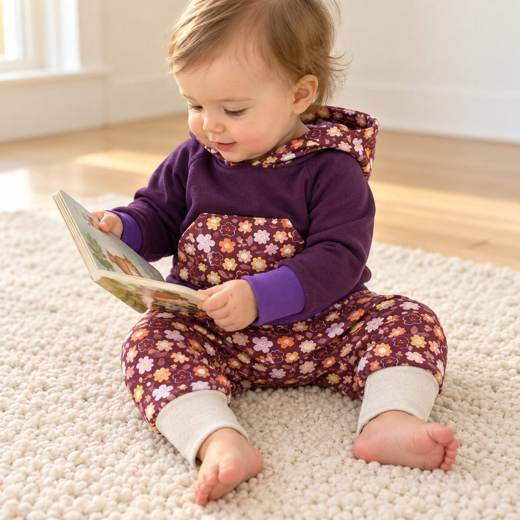Bébé de dix mois feuilletant un livre, vêtu d’un ensemble évolutif avec sweat violet et sarouel à fleurs bordeaux.