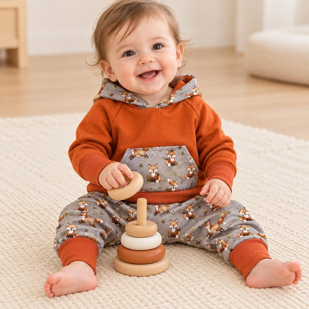 Bébé de dix mois jouant avec des anneaux, portant un sweat terracota à capuche et un sarouel gris à renards.
