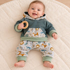 Bébé de six mois tenant un anneau, vêtu d’un sweat vert et d’un sarouel gris à animaux, avec bords sauge repliés.