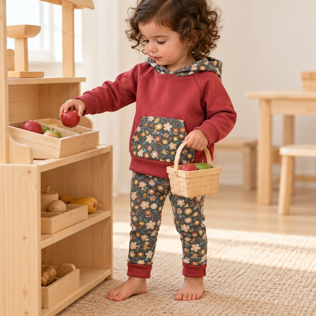 Enfant de deux ans jouant à la marchande, portant un sweat rouge brique et un legging à fleurs sur fond foncé, bords repliés.
