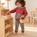 Enfant de deux ans jouant à la marchande, portant un sweat rouge brique et un legging à fleurs sur fond foncé, bords repliés.