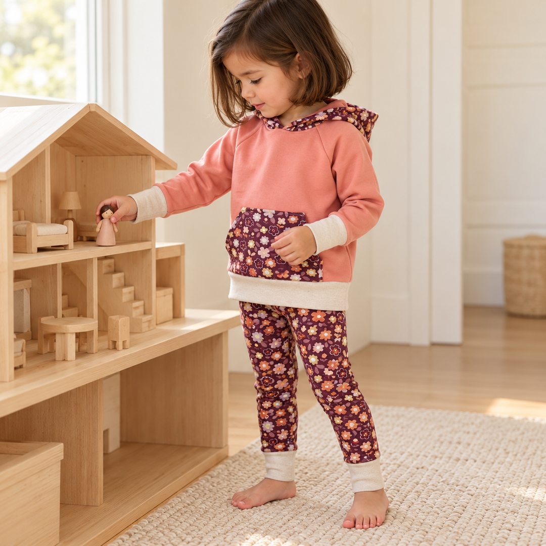 Fille de trois ans jouant avec une maison de poupées, en sweat rose et legging fleuri bordeaux, tous les bords dépliés.