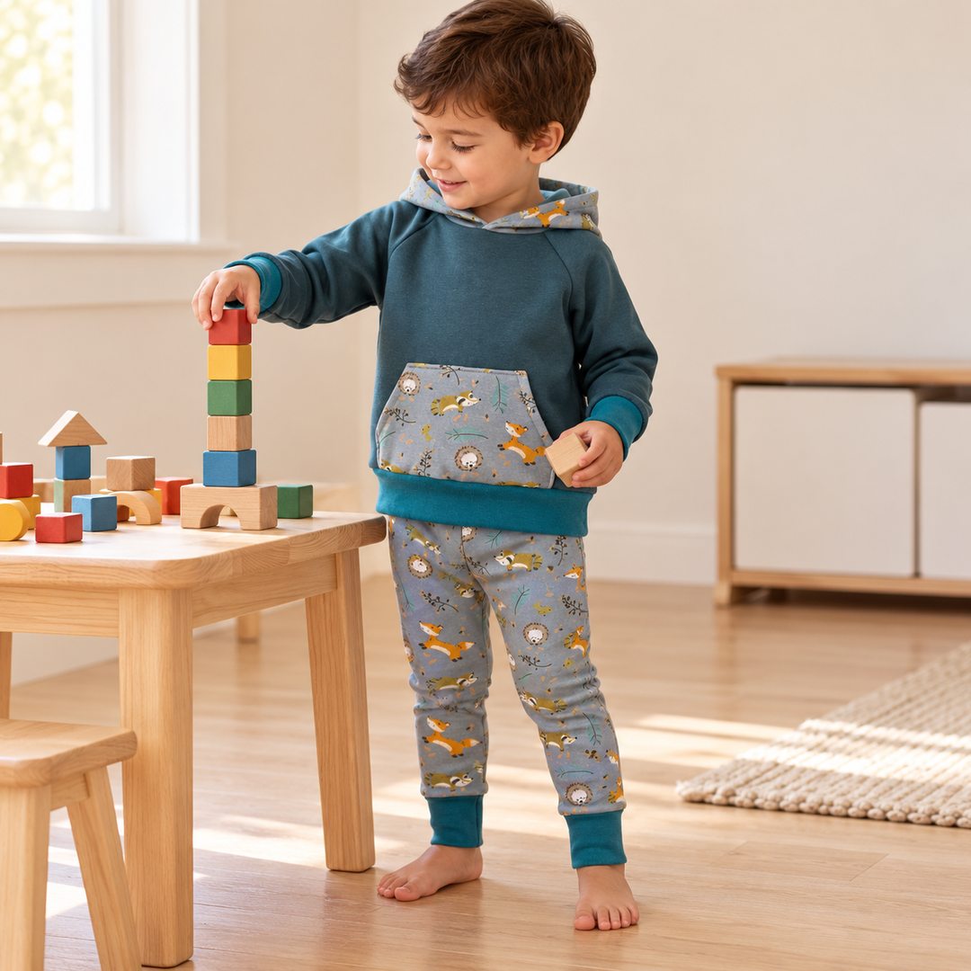 Garçon de trois ans empilant des cubes, en sweat bleu canard et legging gris à animaux de la forêt, bords dépliés.