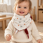 Foulard enfant écru à motifs de champignons et de feuilles, avec doublure rouille foncé, porté par un bébé.