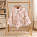 Poncho de siège auto rose à motifs de lapins blancs et de carottes, avec capuche doublée de fourrure douce, suspendu à un portant en bois.